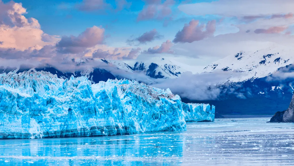 Striking blue glacier floats on calm Alaskan waters. Snow-capped peaks and drifting clouds frame this pristine seascape, showcasing dramatic natural b