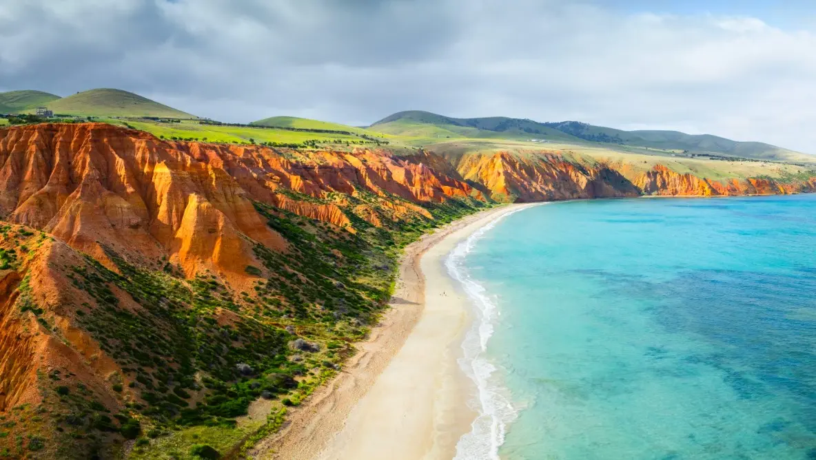 Crystal-blue waves meet a sandy beach along the Australia or New Zealand coast, with green mountains and clear skies defining a serene destination.