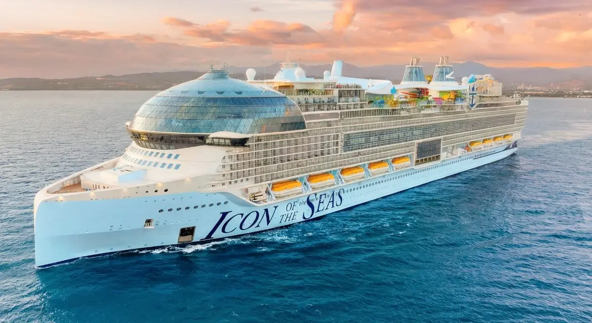 Royal Caribbean International 2026 brand collection: top side view of an Icon-class ship at sea during a vibrant sunset. Experience 2026 with Royal Caribbean. Enjoy twilight views of the Royal Promenade, multi-generational fun, and thrilling activities for all ages.