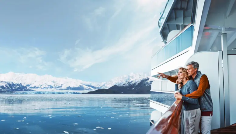 A couple standing on the deck of a Holland America Line ship, admiring a massive blue glacier in Alaska.