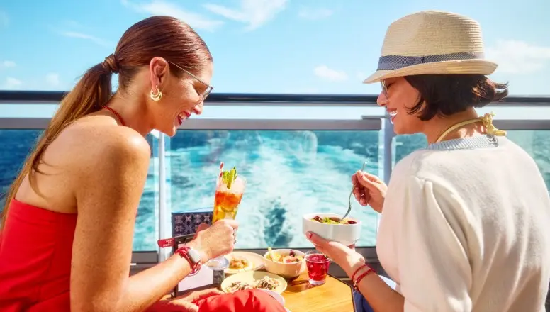 Two stylish women are seated at a table on the sun-drenched deck of a Virgin Voyages cruise ship, enjoying a leisurely brunch with a view of the ocean