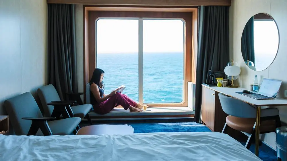Una dama se relaja junto a un gran ventanal en una cabina de crucero con vista al océano, inmersa en un libro cautivador. El ambiente sereno y las impresionantes vistas de la costa crean el refugio perfecto para los huéspedes.