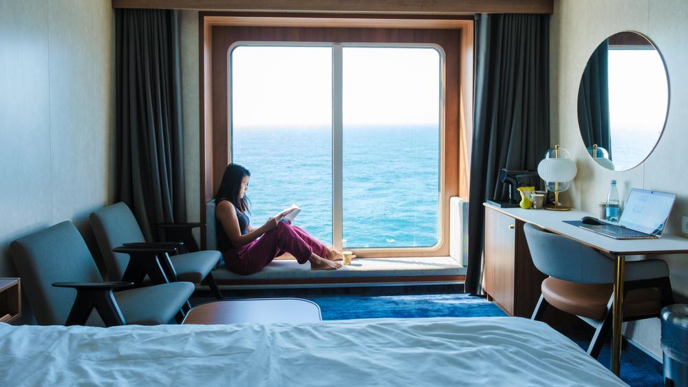 Una dama se relaja junto a un gran ventanal en una cabina de crucero con vista al océano, inmersa en un libro cautivador. El ambiente sereno y las impresionantes vistas de la costa crean el refugio perfecto para los huéspedes.
