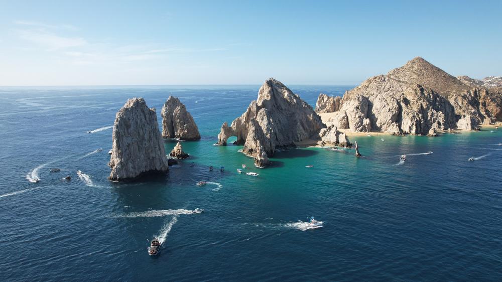 An organized fleet of yachts sailing in the sparkling blue waters of the sea against the backdrop of the famous Cabo San Lucas arch in Mexico