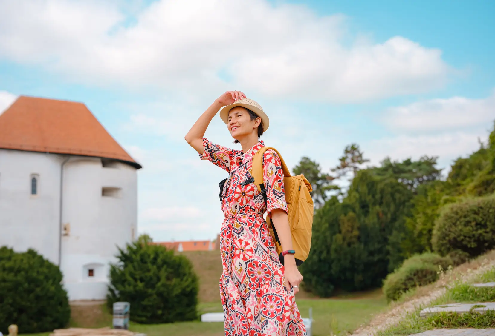 A woman in a vacation-style dress and hat smiles while enjoying the view, wearing a yellow backpack.