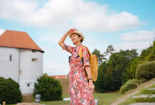 A woman in a vacation-style dress and hat smiles while enjoying the view, wearing a yellow backpack.