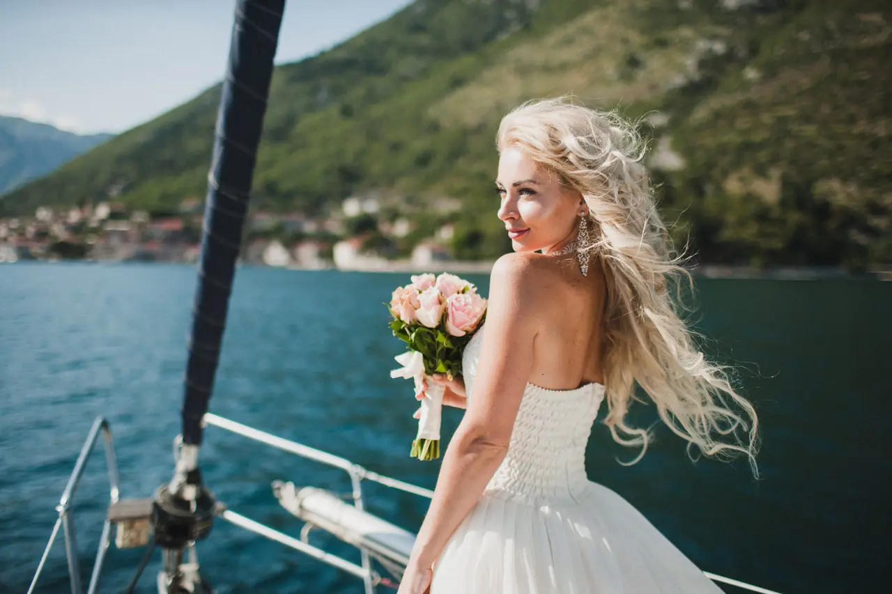 Bride holding a pink rose bouquet, looking back over her shoulder on a cruise deck with a scenic coastal mountain backdrop.