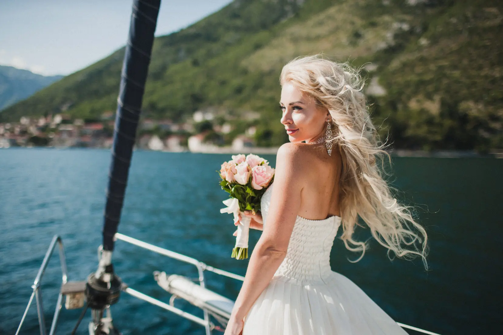 Bride holding a pink rose bouquet, looking back over her shoulder on a cruise deck with a scenic coastal mountain backdrop.