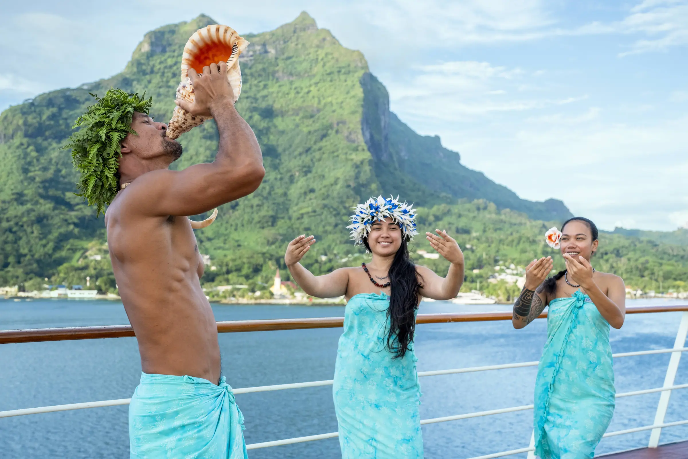 A Les Gauguines performer blows a traditional conch shell as dancers greet guests on deck, with Bora Bora's iconic peak rising in the background.