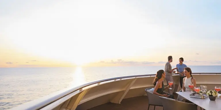 Raise a glass to 35 years of ultra-luxury at sea. Guests gather on an open-air deck aboard a Regent Seven Seas ship as the sun sets over calm ocean waters, a scene that captures the effortless elegance and all-inclusive experience the line has delivered since 1992.