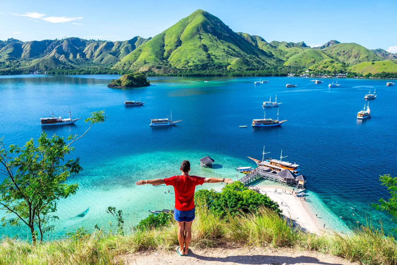 Traveler standing arms wide open on a hilltop overlooking Komodo Island's turquoise bay, with traditional boats anchored below and lush green hills in the background, Indonesia