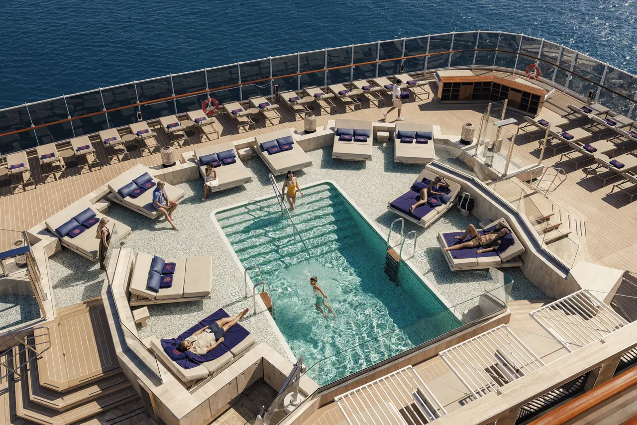 A sun-drenched cruise ship pool deck scene with guests stretched out on cushioned lounge chairs surrounding a calm, blue pool. Passengers enjoy a leisurely day at sea, soaking up the sunshine in a resort-style open-air setting, with the ship's upper decks and railings visible in the background.